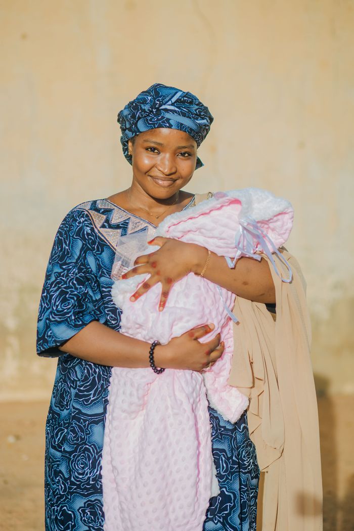 A Nigerian woman joyfully holds her newborn wrapped in pink, wearing traditional attire outdoors.